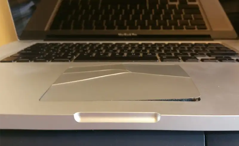 Trackpadul unui MacBook ridicat vizibil din pozitia normala — laptopul nu mai sta plat pe masa, iar trackpadul are colt indoit si fisuri pe margine din cauza bateriei umflate care impinge in sus