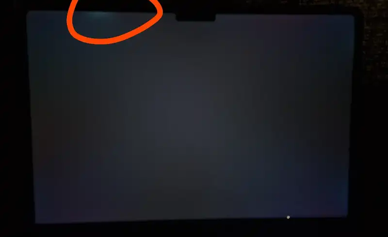 Display MacBook pe ecran negru cu o zona luminoasa evidenta in coltul din stanga-sus incercuita cu portocaliu — backlight bleed sever, semn de panou cu retroiluminare neuniforma sau ramamta in cadre