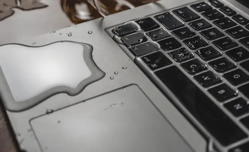 Lichid (apa sau bautura) varsat pe un MacBook — picaturile mari sunt vizibile pe topcase, pe trackpad si in zona tastaturii, exemplu de damage prin infiltrare in mecanismul scissor si pe membrana de sub taste