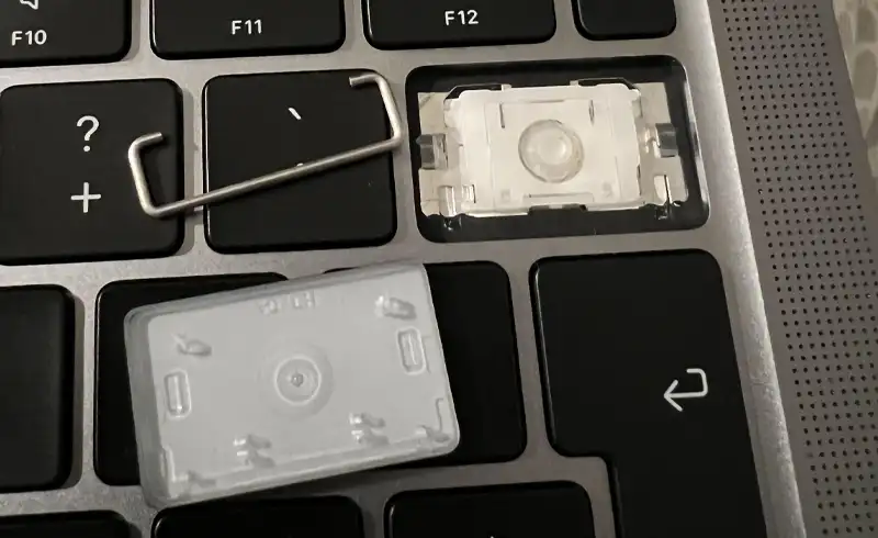 Macro pe tastele F11-F12 ale unei tastaturi MacBook: o tasta lipseste complet expunand mecanismul scissor alb dedesubt, iar keycap-ul scos zace alaturat cu clip-urile metalice vizibile — exemplu de mecanism scissor rupt