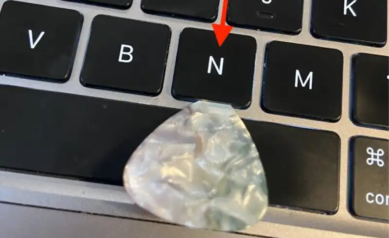 Pick de plastic verde-gri folosit pentru a scoate o tasta blocata pe o tastatura MacBook — sageata rosie indica tasta N care ramane apasata si nu mai revine in pozitia initiala
